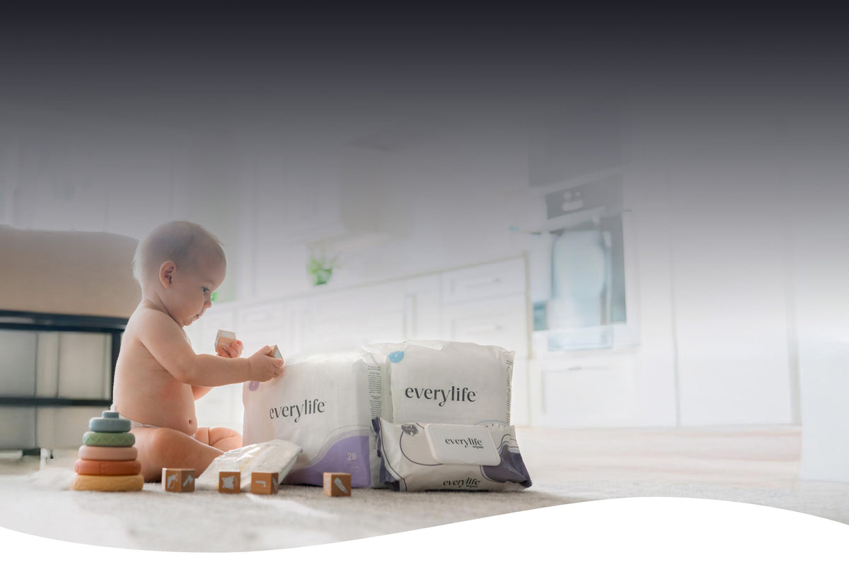 A baby sitting on a floor in a bright room, playing with blocks next to a pack of EveryLife baby wipes and diapers.